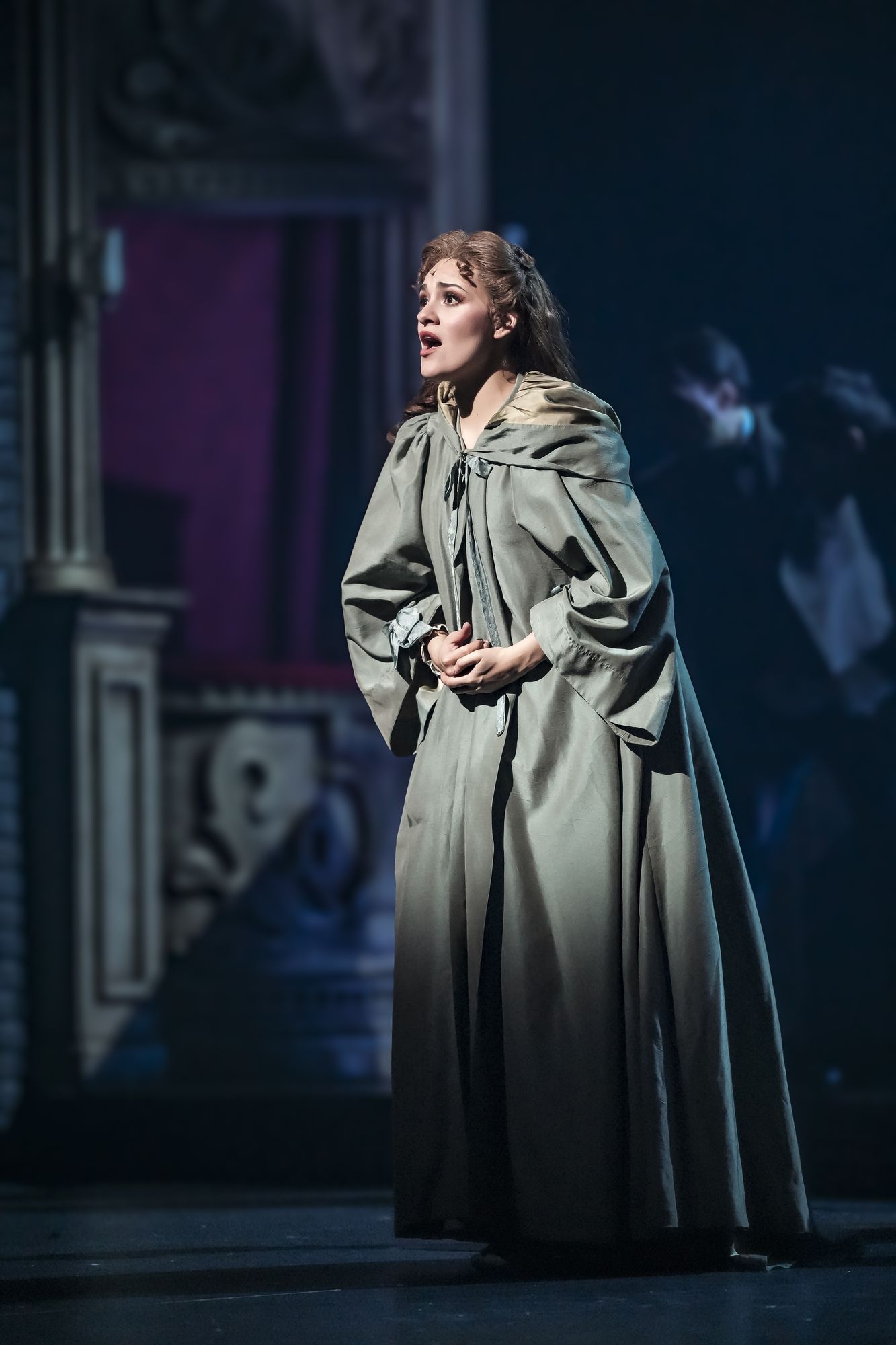 Christine Daaé in a grey travelling gown surrounded by the ensemble