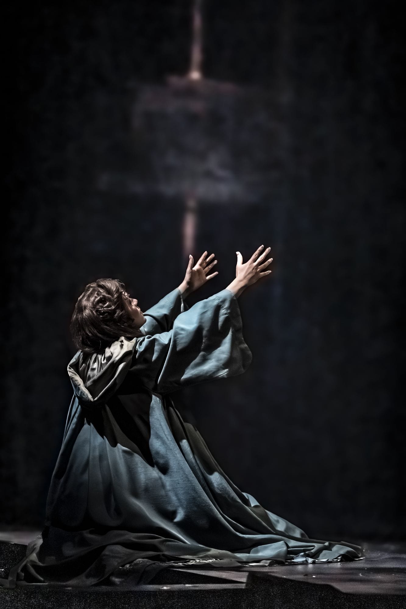 Christine Daaé kneeling with both arms opened wide, lit from above
